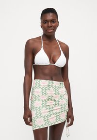 Hvit trekantet bikinioverdel med teksturert stoff, kombinert med en grønn skjørt med rosa blomsterdesign og rynkede detaljer på siden.