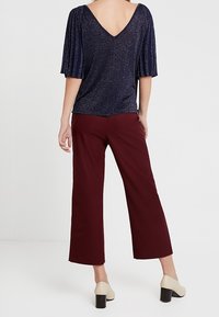 Marineblaues Glitzertop mit kurzen plissierten Ärmeln, tiefem V-Rücken, kombiniert mit bordeauxroten, weit geschnittenen Hosen und cremefarbenen Schuhen mit Blockabsatz.