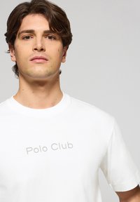 Biała bawełniana koszulka z okrągłym dekoltem i krótkimi rękawami. Posiada lekko szare logo "Polo Club" nadrukowane z przodu.