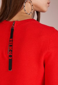 Czerwony sweter z dzianiny z czarnym zamkiem błyskawicznym i logo na uchwycie z tyłu. Duże kolczyki w kształcie kół z koralikowym wzorem.