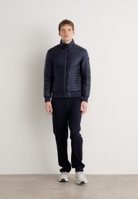 Veste matelassée marine avec des rayures horizontales et un col haut, associée à un pantalon noir et des baskets gris clair. Texture lisse, design ajusté.