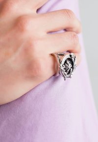 Anillo de plata con un motivo facial detallado, que presenta acentos de hojas en espiral. Lleva en una mano sobre un fondo de tela lavanda.