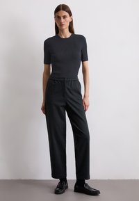 Dark ribbed short-sleeve top paired with high-waisted, loose, dark trousers. Model wears black loafers, standing against a neutral background.