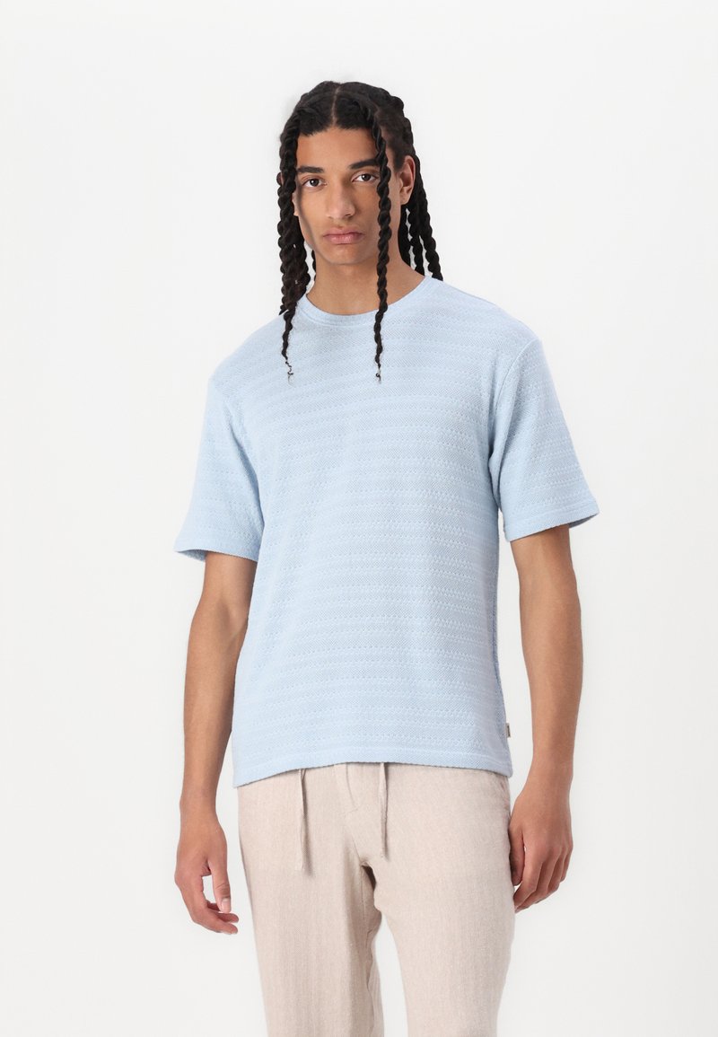 Tricou texturat cu mânecă scurtă de culoare bleu deschis, cu guler rotund și tiv drept, purtat cu pantaloni bej largi.