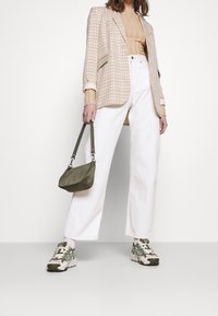 Beige geruite blazer over een geribde tan top, gecombineerd met witte wijde jeans en gepatternde sneakers. Houdt een kleine olijfgroene tas vast.