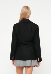 Črn strukturan blejzer s prilegajočim krojem, velikim ovratnikom in gumbnimi detajli na rokavih. Nosijo ga nad svetlo sivo plisirano krilo.