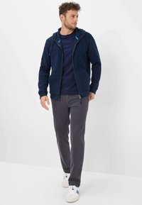 Marineblå lynlås hoodie med blå detaljer, der har en blød tekstur, frontlommer og hætte, parret med mørkegrå bukser og hvide sneakers.