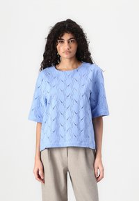 Blauw kortmouwtop met golfpatroon in de oogjes. Vierkante halslijn en een relaxte fit. Gecombineerd met lichtbeige broek.