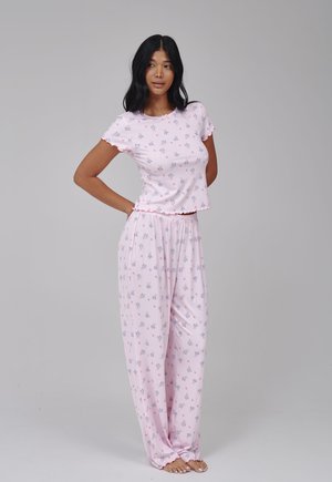 Vrouw staat blootsvoets, draagt een lichtroze shirt met korte mouwen met bloemenprint en losse pyjamabroek, tegen een effen grijze achtergrond.
