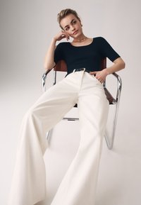 Top negro de canalé con mangas cortas, pantalones de pierna ancha crema y un cinturón plateado, sentado en una silla de cromo con acentos de cuero marrón.