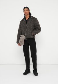Braune strukturierte Jacke mit Reißverschluss, kombiniert mit schwarzen Hosen und einer grauen Tasche. Das Modell trägt schwarze Schuhe und ein Hemd mit Kragen darunter.