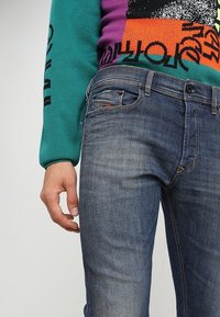 Närbild av person klädd i blå urblekta jeans och en färgglad tröja med mönster i teal, orange, svart och lila, stående med vänster hand avslappnad.