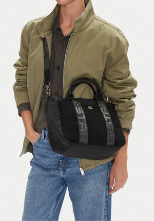 Osoba ubrana w oliwkową kurtkę i niebieskie dżinsy niesie czarną torebkę na ramię i do ręki marki G-STAR przewieszoną przez tors.