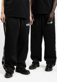 Pantalones joggers anchos negros con un detalle de logo blanco y rayas laterales, hechos de un material suave. Combinados con zapatos negros de suela gruesa.