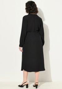 Vestido midi negro con mangas largas, cintura ceñida y tela suave. Presenta una silueta fluida y un sutil detalle de costura en la parte posterior.