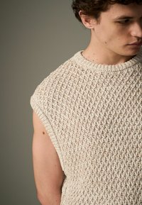 Top sans manches tricoté de couleur beige clair, présentant un motif texturé avec de petites formes de diamant et une finition côtelée au niveau du cou.