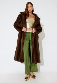 Bruine nepbontjas, gouden glanzende crop top, groene satijnen wijde broek en gouden hakken met strikken. Gladde textuur en oversized ontwerp.