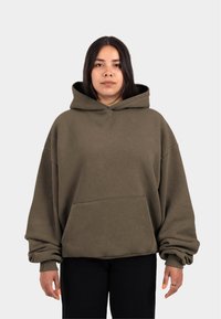Kapuzensweatshirt aus olivgrünem Stoff, mit einer Fronttasche, weiten Ärmeln und einer lockeren Passform. Modell steht vor einem neutralen Hintergrund.
