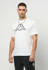 Mann med kort hår iført hvit t-skjorte med svart motiv av to sittende figurer rygg mot rygg, svarte bukser med hvit logo på låret.