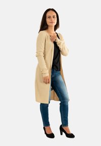 Donna in piedi con un lungo cardigan beige, top nero, jeans blu e tacchi neri su uno sfondo bianco semplice.