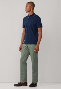 Polo bleu marine avec col blanc et bordure aux manches, associé à un pantalon vert clair et des mocassins marron foncé, posé devant un fond neutre.