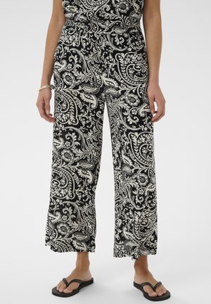 Kvinde iført sorte og hvide bredbenede bukser med paisley-mønster og elastisk talje samt sorte flip-flop sandaler, stående foran en ensfarvet baggrund.