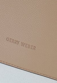 Brauntexturiertes Material mit geprägtem "GERRY WEBER"-Logo, über einem glatten cremefarbenen Abschnitt mit sichtbarem Steppdetail.