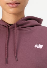 Sudadera burdeos con cordones trenzados, con un logo "NB" en rosa claro en el lado izquierdo del pecho. Está hecha de una tela suave y texturada.