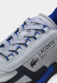 Άσπρο αθλητικό παπούτσι Lacoste με μπλε λεπτομέρειες, λεπτομερείς ραφές, υφή στη σόλα και λογότυπο με κροκόδειλο και κείμενο στην πλαϊνή πλευρά.