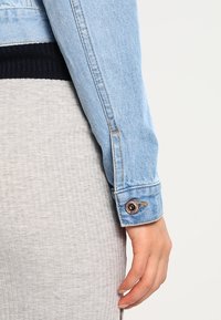 Svetle modrá džínsová bunda se rukávem s knoflíkovým uzávěrem, podrobným prošíváním a ribbed tmavě námořnickým manžetou, v kombinaci se šedou texturovanou tkaninou pod ní.
