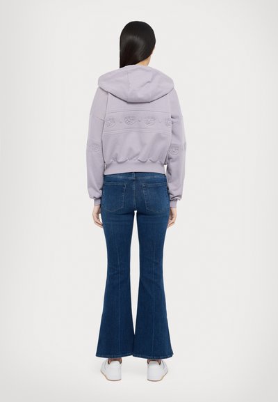 Sweatshirt com capuz lavanda com padrão texturizado, combinado com jeans de boca de sino azul escuro. Ténis brancos completam o visual.