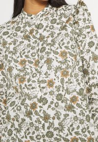 Blouse fleurie avec des motifs verts, orange et blancs. Présente un col avec boutons. Tissu léger avec légère texture.