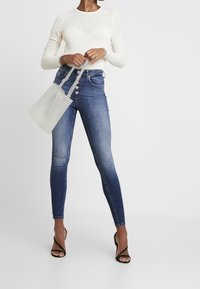 Högmidjade blå denim skinny jeans med en blekningseffekt, tillsammans med en krämfärgad ribbad långärmad topp och en transparent pärlväska.