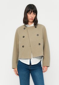 Kvinne iført en beige kort ermet dobbelspent jakke over en hvit skjorte med blå jeans, stående foran en enkel hvit bakgrunn.