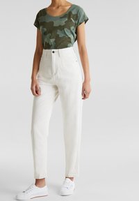 Camiseta verde de camuflaje de manga corta, combinada con jeans blancos de cintura alta y zapatillas blancas, destacando un estilo casual y moderno.