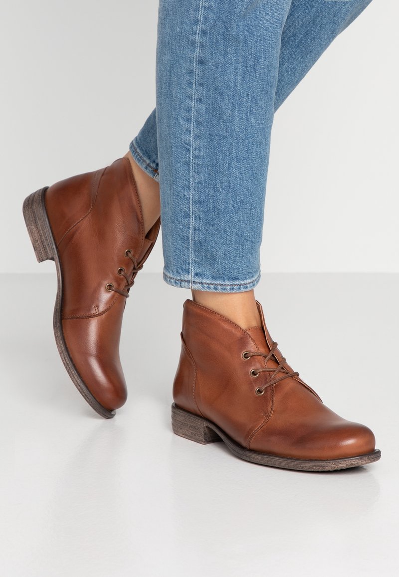 Stivaletti in pelle marrone con lacci indossati con jeans in denim blu su un pavimento bianco.