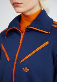 Marinblå zip-jacka med oranga dragkedjor och accentränder, med en texturerad tyg och lilla broderad logotyp på bröstet.