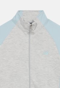 Svetlosivá bunda na zips so svetlomodrými rukávmi, vyrobená z mäkkej tkaniny. Má vysoký golier a malé logo na hrudi.