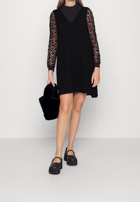 Robe en tricot noir avec un décolleté en V, manches longues transparentes avec un motif en forme de cœur, associée à des chaussures à plateforme noires et un sac en fausse fourrure noir.