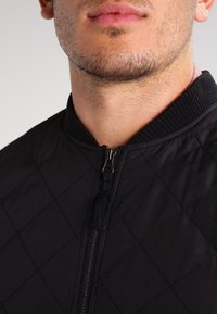 Nahaufnahme des unteren Gesichts und Halses eines Mannes, der eine schwarze gesteppte Jacke mit einem Reißverschluss vorne und einem gerippten Kragen trägt.