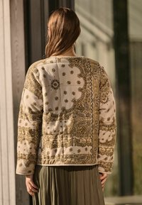 Gequilte jas in crème met ingewikkelde gouden en bruine paisley patronen. Heeft een relaxte pasvorm, lange mouwen en een cropped zoom.