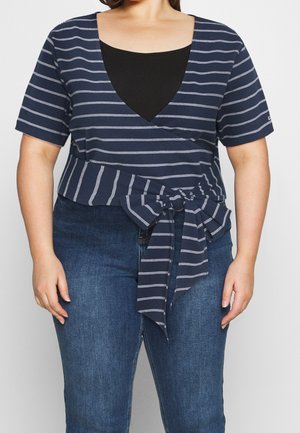 Kvinde iført en navyblå og hvide stribet omslagstop med sløjfe bundet i taljen over en sort undertrøje og blå jeans.