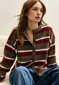 Cardigan rayé en marron, bordeaux et blanc. Présente un col rond, une fermeture à boutons et une texture douce. Assorti avec un denim bleu ample.