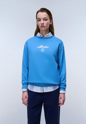 Jonge vrouw die een blauwe Napapijri-trui draagt over een gestreept overhemd en donkere broek, staand tegen een effen lichte achtergrond.