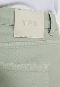 Los pantalones de mezclilla verde claro presentan una tela texturizada, un parche rectangular de cuero crema con el grabado "YPS", y costuras limpias a lo largo de los bolsillos.