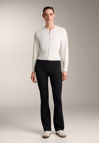 Bela prilegajoča jopica z rebrasto teksturo in zadrgo, v kombinaciji s črnimi širokimi hlačami in belimi superge, ki imajo bež poudarke.