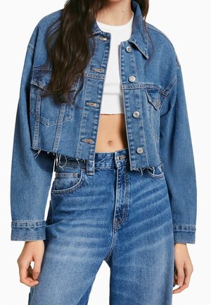 Kvinde iført cropped blå denimjakke med sølvknapper, blå jeans med høj talje og hvid cropped top, der viser maven.