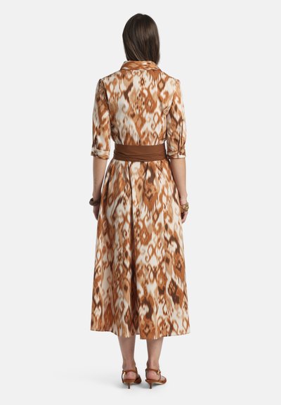 Frau trägt ein braun-beiges gemustertes Midikleid mit dreiviertel Ärmeln und einem breiten braunen Gürtel, steht mit dem Rücken zur Kamera vor einem weißen Hintergrund.