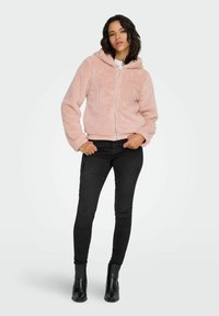 Roza, puhast zip-up majica s kapuco, rebrastimi manšeti in robom. Združena s črnimi ozkimi hlačami in črnimi gležnjaki. Gladek tekstura.