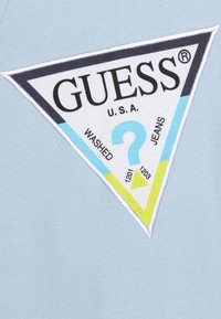Světle modrá látka má velké trojúhelníkové logo v černé, modré a zelené barvě s tučným textem "GUESS," "WASHED," a "JEANS."